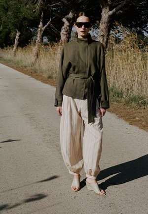 Mujer con chaqueta verde oscuro con cinturón, pantalones holgados a rayas, sandalias beige de cuña y gafas de sol, de pie en un camino soleado junto a hierba alta.