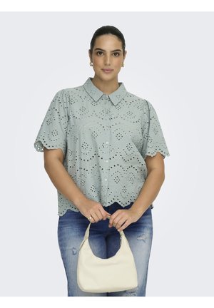 Femme portant un chemisier vert clair à œillets avec des bords festonnés, un jean bleu déchiré, tenant un petit sac à main crème devant un fond uni.