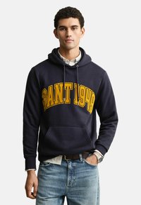 Marineblaues Hoodie mit großen gelben "GANT 1949"-Schriftzug, frontale Kängurutasche, Kapuze mit Kordelzug, gerippte Bündchen, über einem weißen Shirt getragen.