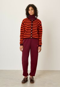 Cardigan rayé orange et noir avec des boutons, porté sur un col roulé bordeaux. Assorti avec un pantalon bordeaux et des chaussures à lacets marron.