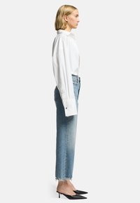 Camicia bianca oversize con bottoni, jeans a vita alta azzurri chiari con orli sfilacciati e décolleté neri a punta. Vista di profilo.