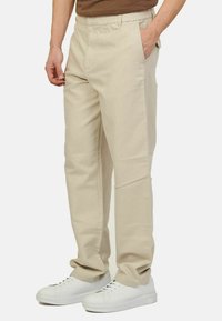 Pantaloni in cotone beige con una vestibilità rilassata, caratterizzati da una parte frontale liscia, tasche laterali e una vita elasticizzata. Abbinati a sneakers bianche.