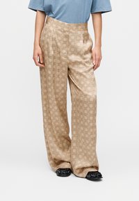 Smėlio spalvos platėjančios kelnės su geometriniu raštu, pasižyminčios lygiu paviršiumi ir aukštu liemeniu, derinamos su juodais batais.