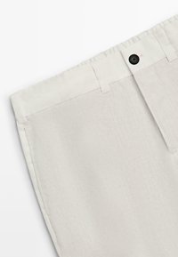 Pantalon en lin beige clair avec taille élastique, passants pour ceinture, poche latérale et fermeture à bouton noir sur le côté droit avant.