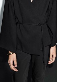 Personne portant un haut enveloppant noir noué à la taille, un pantalon noir et un large bracelet en métal au poignet gauche, contre un fond sombre.
