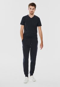 Czarna koszulka z dekoltem w serek w połączeniu z ciemnymi spodniami dresowymi. Spodnie mają boczne kieszenie i mankiety. Model ma na sobie białe trampki. Gładkie tło.