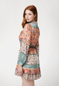 Gėlėmis marginta suknelė, kurioje dera persikų ir žali atspalviai, ilgos rankovės, apykaklės iškirptė ir raukiniai apatinėje dalyje. Tekstūruota medžiaga su sluoksniuotais raštais.