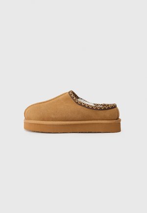 Chaussure slip-on en daim beige avec un bout arrondi, des coutures texturées et une épaisse semelle en caoutchouc beige. Accents tressés décoratifs à l'ouverture.