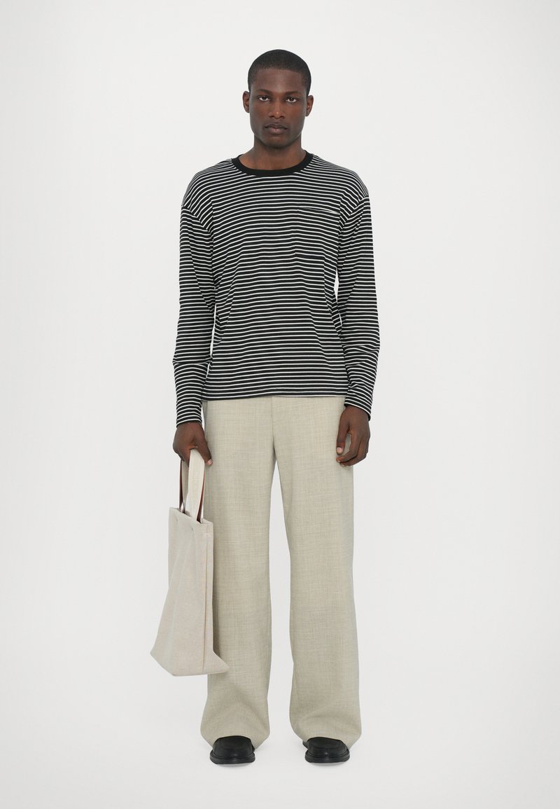 Gestreiftes schwarz-weißes Langarmshirt, hellbeige weite Hose, eine cremefarbene Tragetasche haltend, stehend vor einem schlichten Hintergrund.