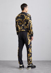 Melns sporta krekls un bikses ar zelta spirāļu rakstu, elastīgiem āmuriem un atvieglotu piegriezumu. Apavi ir melni ar baltiem akcentiem.