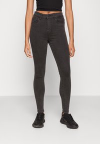 Calças skinny pretas feitas de uma mistura de algodão com uma textura suave, cintura alta, bolsos frontais e passadores de cinto. Combinadas com ténis pretos desportivos.