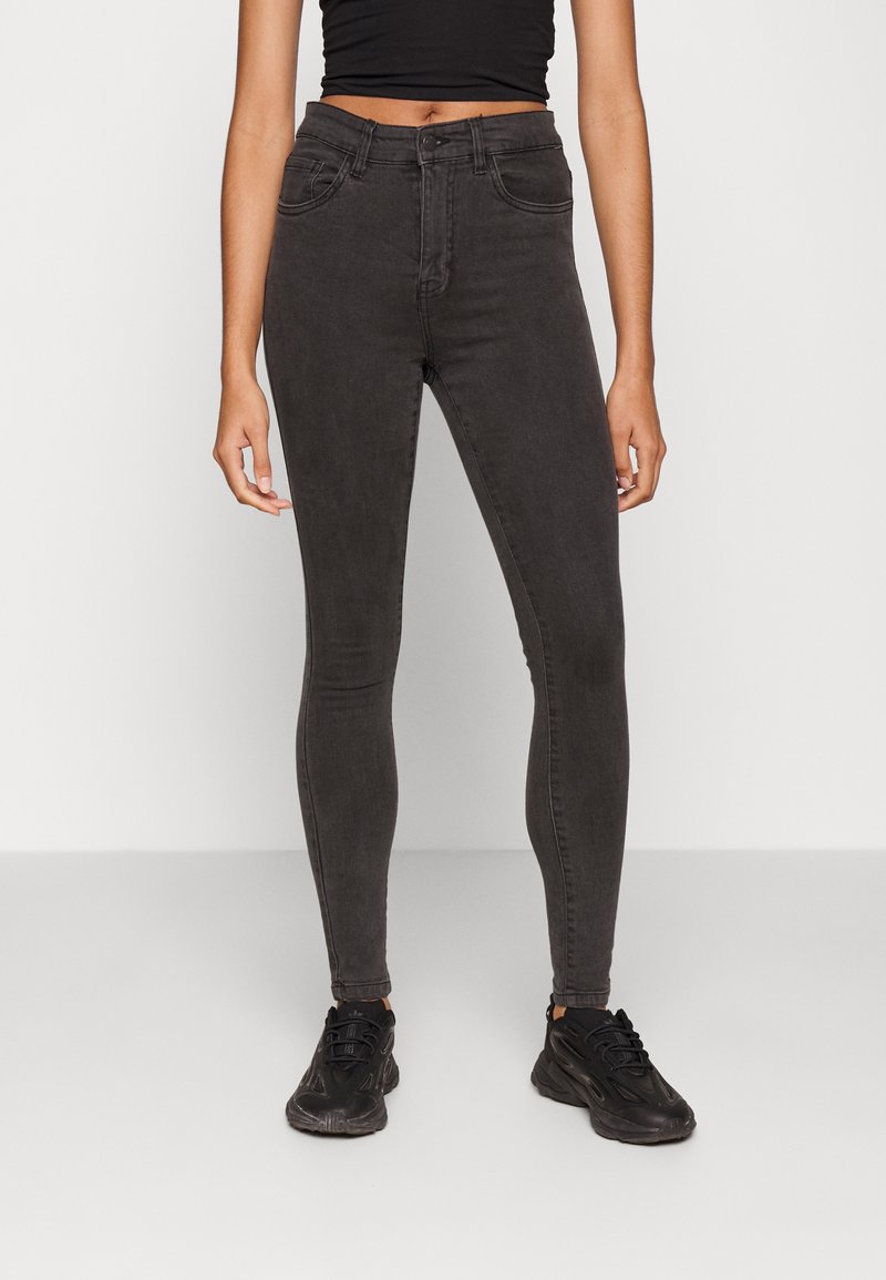 Calças skinny pretas feitas de uma mistura de algodão com uma textura suave, cintura alta, bolsos frontais e passadores de cinto. Combinadas com ténis pretos desportivos.