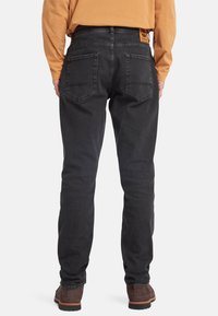 Svarta denimjeans med slim fit, med två bakfickor och en ljusbrun läderlogotyp på midjebandet. Bärs med en långärmad orange skjorta.