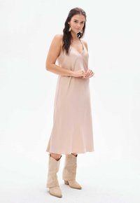 Helles rosa Satin-Kleid mit fließender Silhouette, Spaghetti-Trägern und dezentem Glanz, kombiniert mit beigen Cowboy-Stiefeln. Einfaches Design.