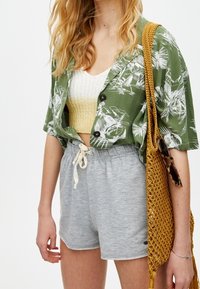 Grüne, mit Blumenmuster versehene, übergroße Bluse, weißes gestricktes Crop-Top, graue Shorts mit Kordelzug und eine gelbe, gewebte Umhängetasche.