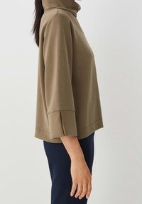 Tan tröja med högt krage och 3/4-långa ärmar som har sprunddetaljer vid ärmsluten, strukturerad design och slät textur. Bärs med mörka byxor.