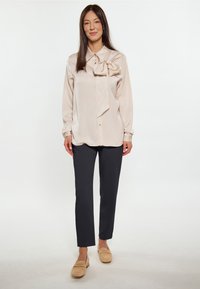 Blusa beige chiaro con dettaglio a fiocco, chiusura a un bottone e maniche lunghe, abbinata a pantaloni blu scuro e scarpe beige.