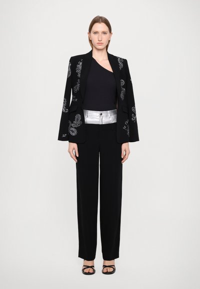 Zwarte blazer met witte paisley-borduring, gecombineerd met een zwarte schouderloze top, zilveren riem en wijde broek. Platte sandalen maken de look compleet.