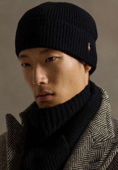 Joven hombre que lleva un gorro de punto negro y bufanda con un abrigo beige de espiga, mirando ligeramente hacia abajo sobre un fondo neutro.