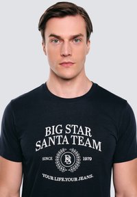 Námořnicky modré bavlněné tričko s bílým textem, kruhovým logem a dekoračními prvky. Krátký rukáv a kulatý výstřih.