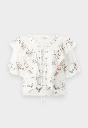 Femme portant un chemisier blanc brodé de fleurs avec des volants, une jupe plissée jaune et des sandales beige avec un nœud, debout avec une main sur la hanche.