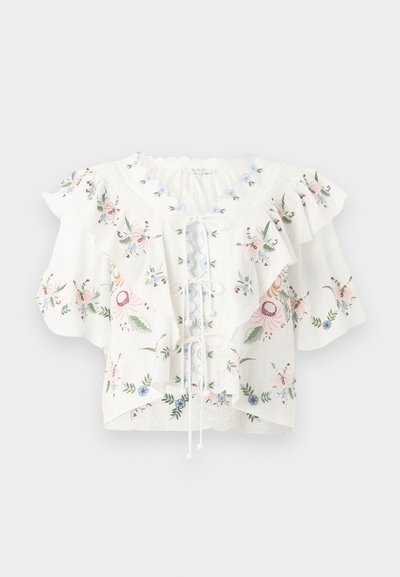 Blouse blanche avec broderie florale, larges manches à volants, encolure festonnée et détail lacé sur le devant.