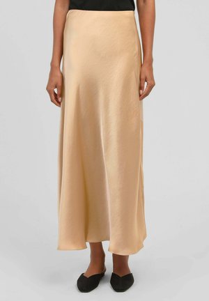 Femme portant une longue jupe beige soyeuse associée à des chaussures plates noires à bout pointu sur un fond clair uni.