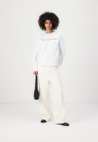 Felpa bianca con il logo "TOMMY HILFIGER", abbinata a jeans bianchi a vita alta. Il modello tiene in mano una borsa nera, con dettagli del logo.