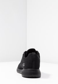 Černá sportovní teniska s texturovanou pleteninou, žebrovanou podrážkou a logem na boku, z pohledu zezadu na bílé ploše.