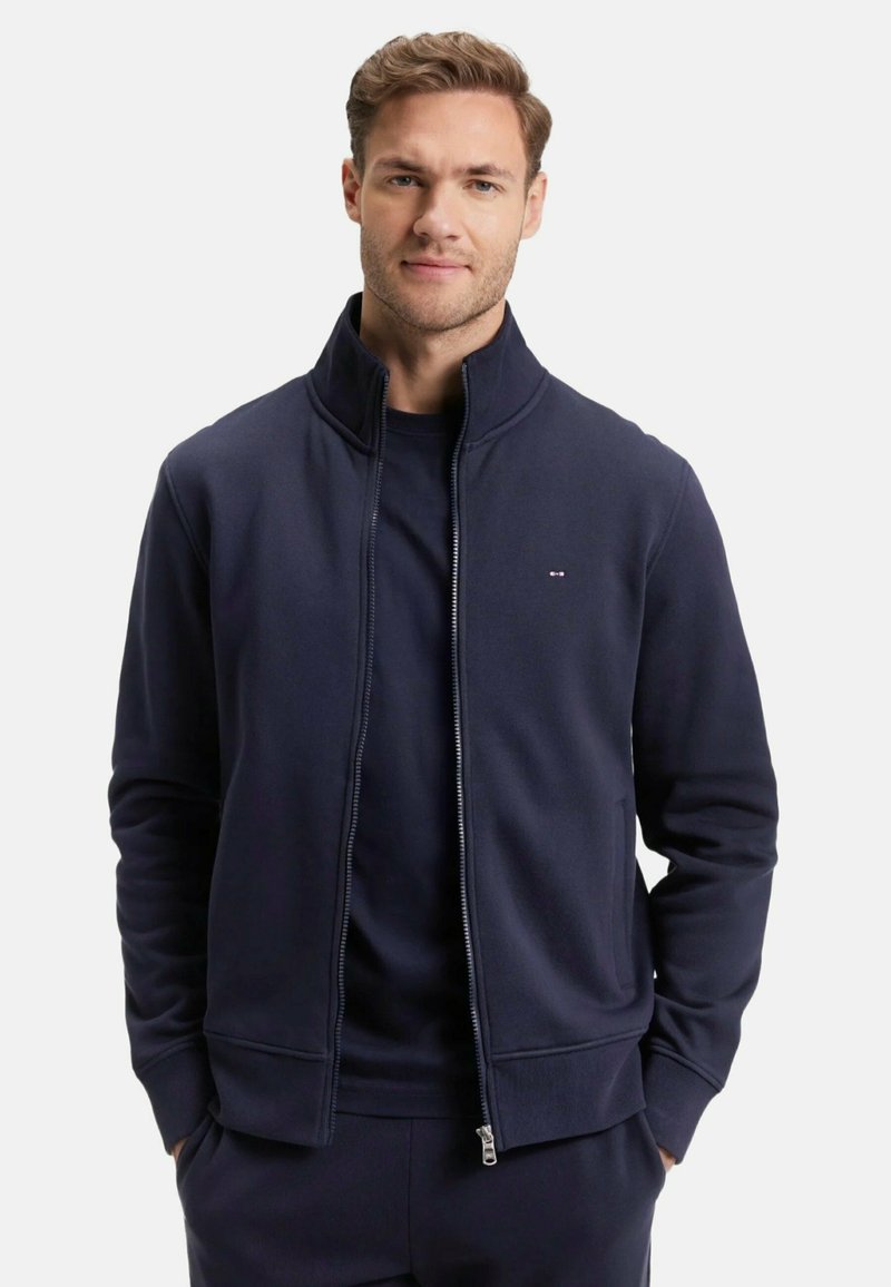 Veste zippée marine avec col haut, fabriquée en tissu doux. Elle possède des poches latérales et un petit logo sur la poitrine. Texture lisse et coupe ajustée.