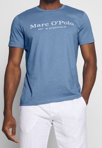 Camiseta de algodón azul con el texto blanco "Marc O'Polo EST. IN STOCKHOLM". Diseño de manga corta y cuello redondo. Textura suave.