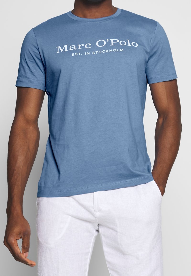 Camiseta de algodón azul con el texto blanco "Marc O'Polo EST. IN STOCKHOLM". Diseño de manga corta y cuello redondo. Textura suave.