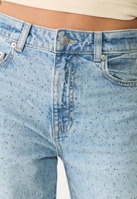 Jeans de denim azul claro con adornos metálicos dispersos. Diseño clásico con cierre de botón y bolsillos frontales.