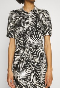 Femme portant une robe noire et blanche à motifs de feuilles avec des boutons, des manches courtes et une ceinture à nouer à la taille.
