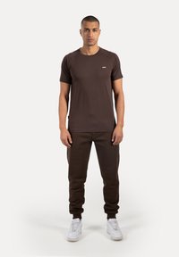 Brun kortærmet t-shirt med rund halsudskæring og et lille logo, kombineret med mørkebrune joggers og hvide sneakers.