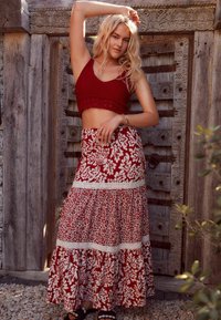 Blond kvinde i rød hæklede crop top og lang rød blomsterkjole poserer med den ene arm hævet foran en baggrund af udskåret trækdør.