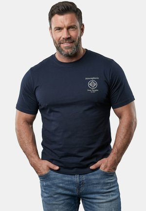 Man met baard die een marineblauw T-shirt met de tekst "Atmospheric Deep Thoughts Est. 2007" draagt en een blauwe spijkerbroek, handen in de zakken, glimlachend naar de camera.