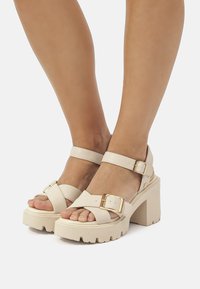 Beige Plattformsandalen mit Blockabsatz, verstellbaren Riemen, metallischen Schnallen, strukturiertem Fußbett und glattem lederähnlichem Material.