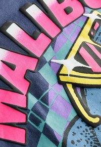 Grafika s tučným růžovým textem "Malibu," umístěným na vícerozměrné geometrické vzoru v modré, zelené a fialové barvě se strukturovanými akcenty.