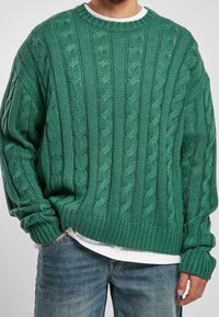 Pull vert en maille avec un motif torsadé, col rond, poignets et ourlet côtelés. Porté par-dessus une chemise blanche, avec une coupe décontractée.