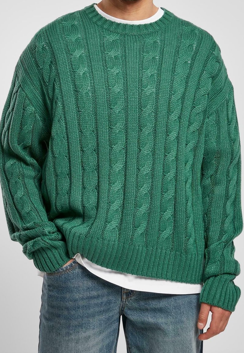 Pull vert en maille avec un motif torsadé, col rond, poignets et ourlet côtelés. Porté par-dessus une chemise blanche, avec une coupe décontractée.