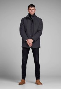 Pelēks vilnas mētelis ar augstu apkakli un trim pogām, kombinēts ar melnām šaurām džinsiem un brūniem zābakiem līdz potītei. Gluda struktūra, minimālistisks dizains.