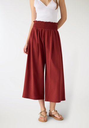 Donna che indossa pantaloni a vita alta, a gamba larga, di colore rosso ruggine con una fascia arricciata in vita, abbinati a un top bianco senza maniche e sandali color cuoio.
