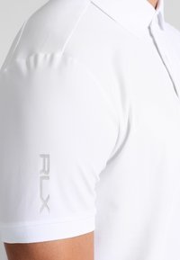 Bílá polo tričko vyrobené z hladké tkaniny, s límečkem, krátkými rukávy a stříbrným logem "RLX" na levém rukávu.