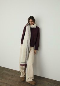 Maglione in maglia bordeaux, sciarpa crema con righe bordeaux, pantaloni chiari a campana beige, pantofole in suede marrone, sfondo in legno.