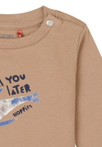 Chemise à manches longues beige avec col rond. Présente des boutons-pression en métal à l'épaule et un imprimé graphique bleu avec du texte sur le devant.