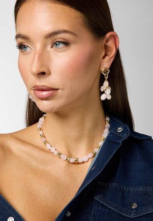 Kobieta o prostych brązowych włosach, nosząca jasnoróżowy naszyjnik z koralików i pasujące wiszące kolczyki, ubrana w ciemnoniebieską bluzkę jeansową.