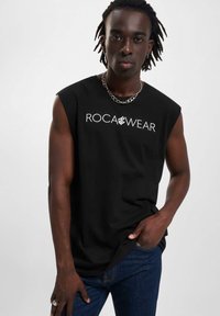 Černá tílko bez rukávů s bílým logem "ROCA WEAR" kombinované s tmavě modrými džíny. Model má krátké, kudrnaté vlasy a řetízkový náhrdelník.