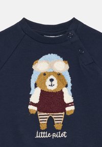 Marinblå sweatshirt med en taktil björn-design iförd en blå pilotmössa, vinröda byxor och randiga långbyxor. Texten "Liten pilot" längst ner.