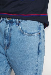 Ljusblå denimjeans med en klassisk passform, med en framficka och kontrasterande orange sömmar. Matchas med en marinblå skjorta med röda och blå ränder.